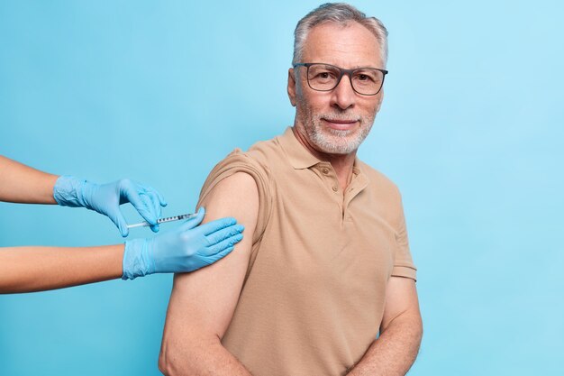 Bearded grey haired senior man gets vaccination against coronavirus protects himself from virus wears spectacles and t shirt looks determined isolated over blue wall