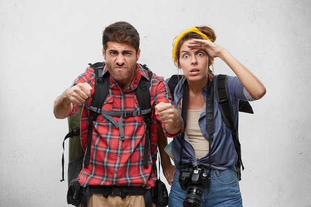 Bearded guy in plaid shirt with backpack driving car, holding tight driving wheel while his girlfriend next to him keeping hand on her forehead, staring into distance with shocked amazed look