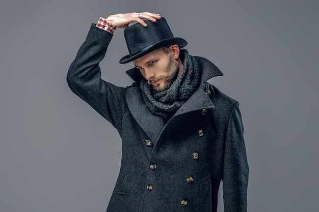 Free photo a bearded hipster male dressed in a gray jacket and a cylinder hat isolated on grey background.