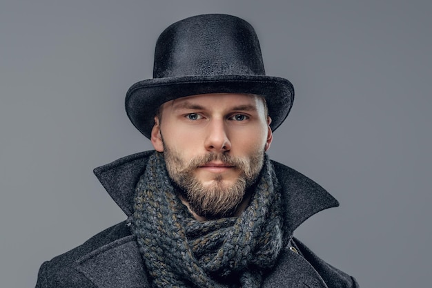 Free photo a bearded hipster male dressed in a gray jacket and a cylinder hat isolated on grey background.