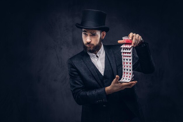 A Bearded magician in a black suit and top hat, showing trick with playing cards on a dark background.