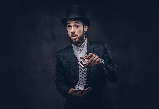 Free photo a bearded magician in a black suit and top hat, showing trick with playing cards on a dark background.