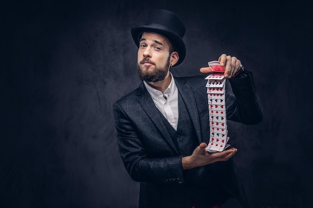 Free photo a bearded magician in a black suit and top hat, showing trick with playing cards on a dark background.