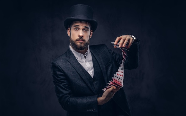 A Bearded magician in a black suit and top hat, showing trick with playing cards on a dark background.