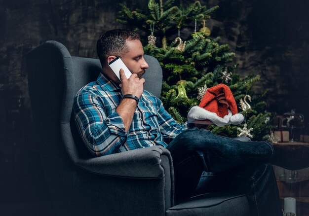 Bearded male dressed in a blue plaid shirt sits on a chair and talks by smartphone over Christmas decorated background.