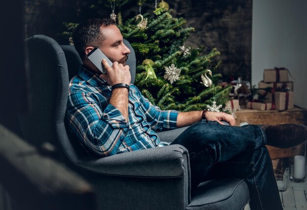 Bearded male dressed in a blue plaid shirt sits on a chair and talks by smartphone over Christmas decorated background.