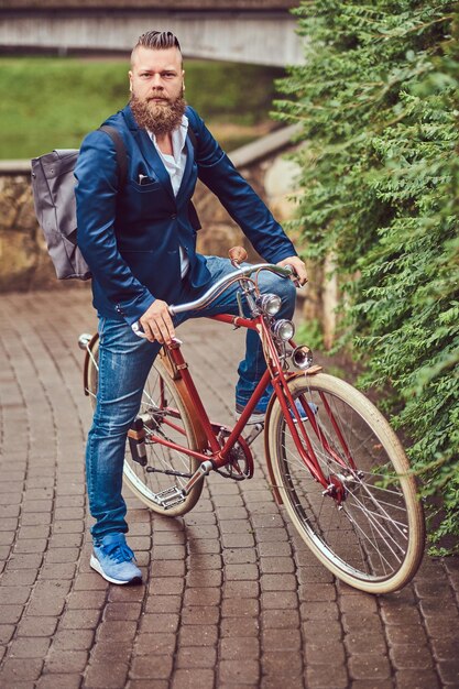 Bearded male with a stylish haircut dressed in casual clothes with a backpack, sitting on a retro bicycle in a city park.