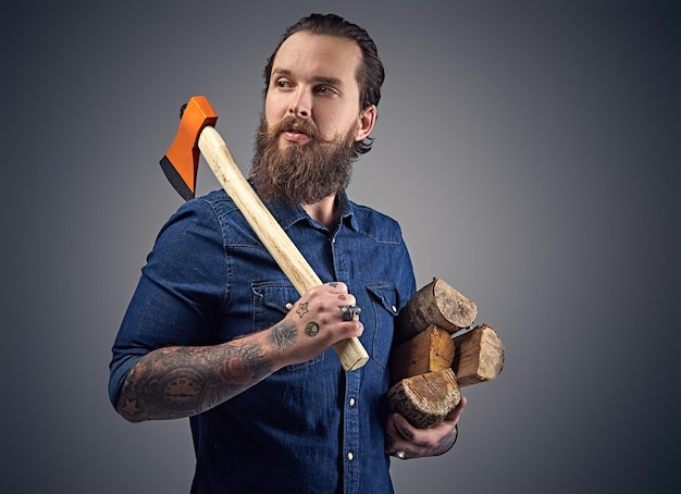 Free photo bearded male with tattoos on arms wearing a denim shirt holds an axe and firewoods over grey background.