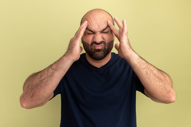 Bearded man in black t-shirt looking unwell and annoyed touching his head suffering from strong headache standing over green wall