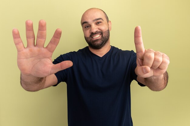 Bearded man in black t-shirt  smiling showing number six standing over green wall