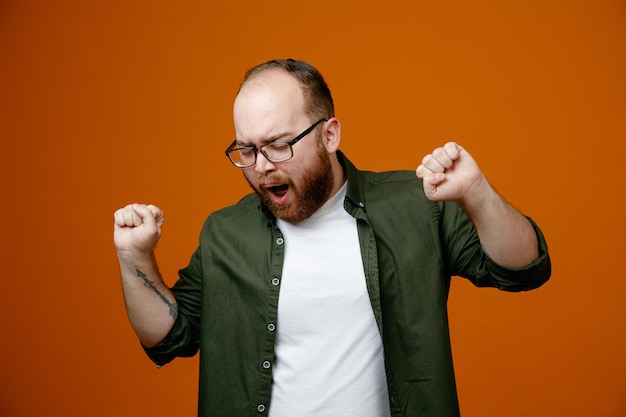 Bearded man in casual clothes clenching fists crazy happy rejoicing his success standing over orange background