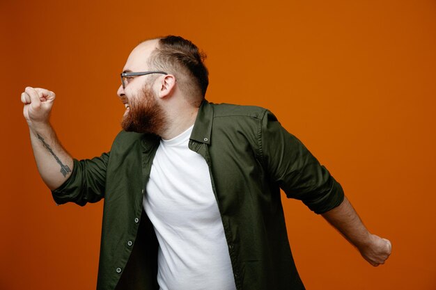 Bearded man in casual clothes clenching fists crazy happy rejoicing his success standing over orange background