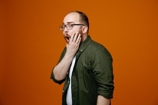 Bearded man in casual clothes looking at camera amazed and surprised covering mouth with hand standing over orange background