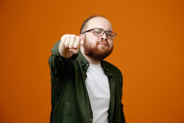 Free photo bearded man in casual clothes wearing glasses clenching fist pointing it at cakmera looking confident standing over orange background