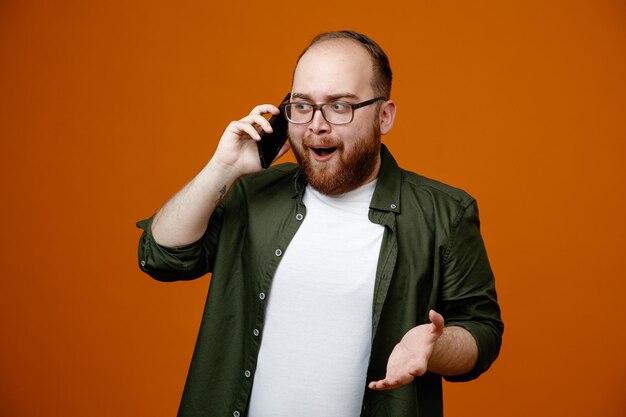 Bearded man in casual clothes wearing glasses happy and surprised smiling while talking on mobile phone standing over orange background