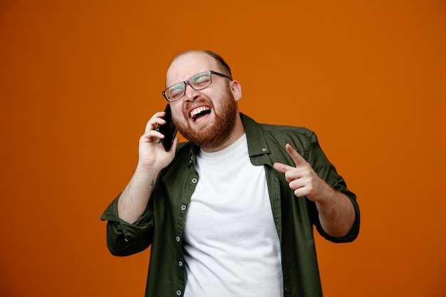 Bearded man in casual clothes wearing glasses laughing out while talking on mobile phone standing over orange background