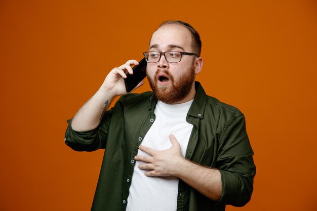 Bearded man in casual clothes wearing glasses looking confused while talking on mobile phone standing over orange background