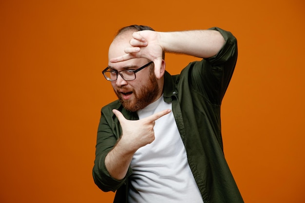 Bearded man in casual clothes wearing glasses making frame with fingers looking at camera through this frame happy and cheerful standing over orange background