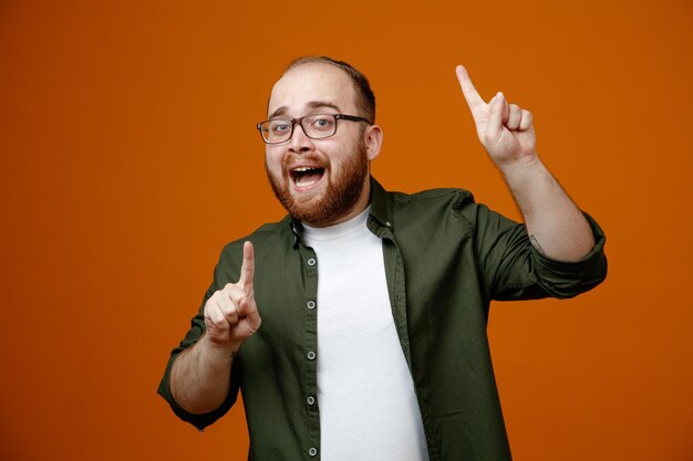 Bearded man in casual clothes wearing glasses pointing with index fingers up happy and pleased smiling cheerfully standing over orange background