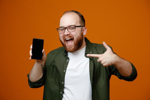 Bearded man in casual clothes wearing glasses showing smartphone pointing with index finger at it smiling cheerfully standing over orange background