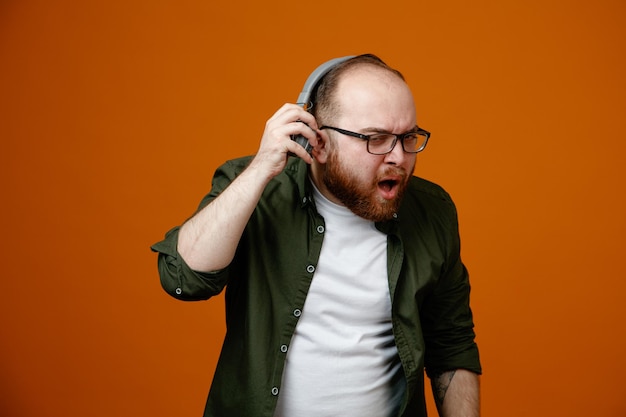 Bearded man in casual clothes wearing glasses with headphones looking at camera being confused trying to hear better taking off headphones standing over orange background