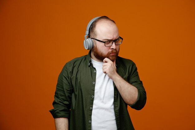 Bearded man in casual clothes wearing glasses with headphones looking at camera with pensive expression thinking holding hand on his chin standing over orange background