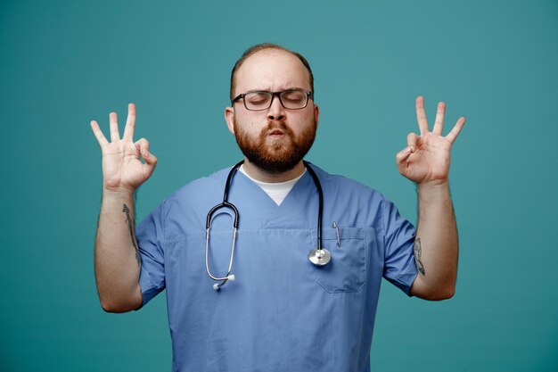 Bearded man doctor in uniform with stethoscope around neck wearing glasses meditating making meditation gesture with fingers calm and relaxed standing over blue background