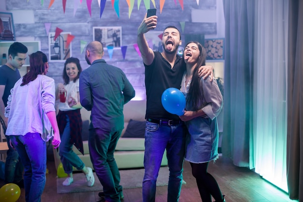 Free photo bearded man and his girlfriend with tongue out taking a selfie while partying with their friends.