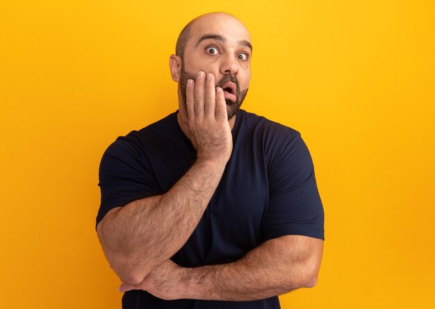 Bearded man in navy t-shirt  amazed and surprised standing over orange wall