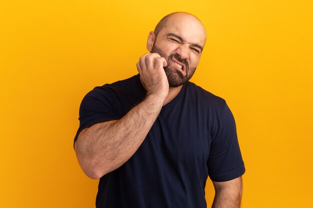 Bearded man in navy t-shirt looking confused scratching his face standing over orange wall