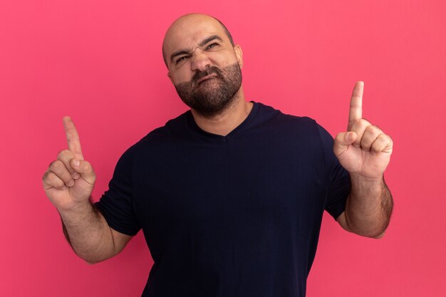 Bearded man in navy t-shirt looking up making wry mouth with disappointed expression showing index fingers standing over pink wall