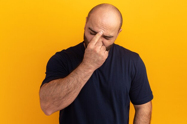 Bearded man in navy t-shirt touching his forehead tired and bored having headache standing over orange wall