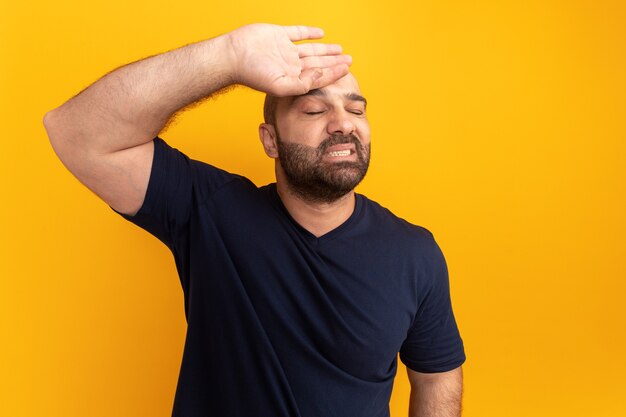 Bearded man in navy t-shirt with hand on his forehead with annoyed expression standing over orange wall