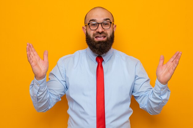 Free photo bearded man in red tie and blue shirt wearing glasses looking at camera happy and positive smiling cheerfully raising hands standing over orange background