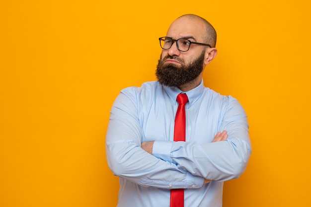 Free photo bearded man in red tie and shirt wearing glasses looking aside with arms crossed with skeptic expression