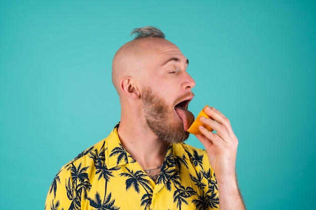 Free Photo a bearded man in a shirt with a print of palm trees on a turquoise wall licks an orange