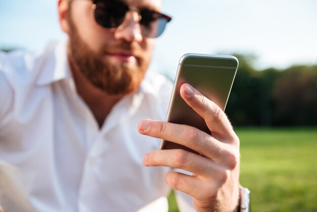 bearded man in sunglasses and shirt while using smartphone. Focus on phone