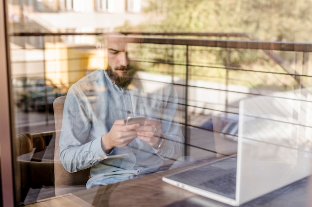 Free photo bearded man using mobile phone seen through transparent glass in caf�