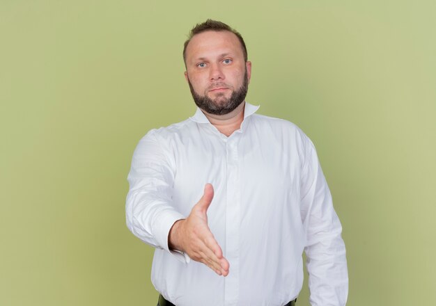 Bearded man wearing white shirt greeting offering hand standing over light wall