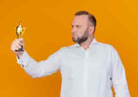 Free photo bearded man wearing white shirt holding trophy looking at it with serious face standing over orange wall