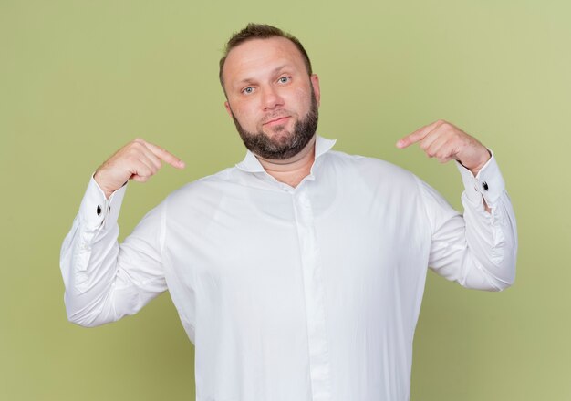 Free photo bearded man wearing white shirt looking confident pointing with fingers at himself standing over light wall