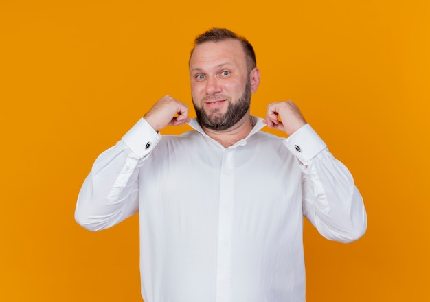 Bearded man wearing white shirt  smiling confident fixing his collar standing over orange wall