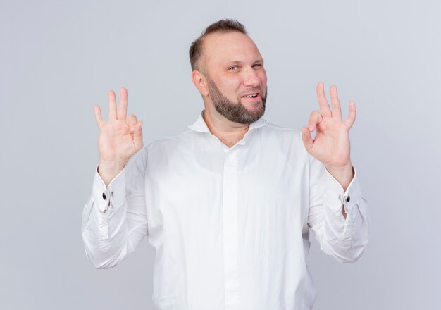 Bearded man wearing white shirt  smiling confident showing ok sign standing over white wall