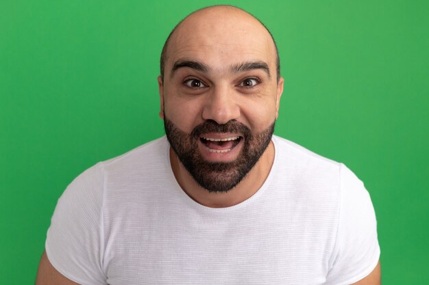 Bearded man in white t-shirt  happy and excited smiling cheerfully standing over green wall