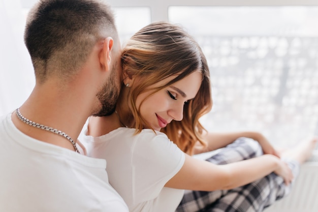 Bearded man in white t-shirt kissing girlfriend's hair during indoor photoshoot. Portrait of winsome curly blonde woman embracing with husband.