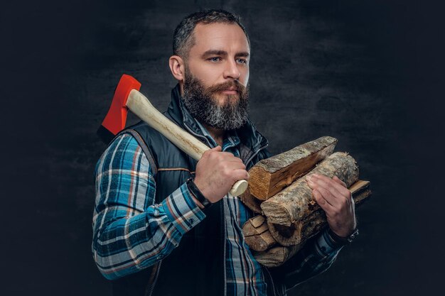 Bearded middle age male holds an axe and firewoods over dark grey background.