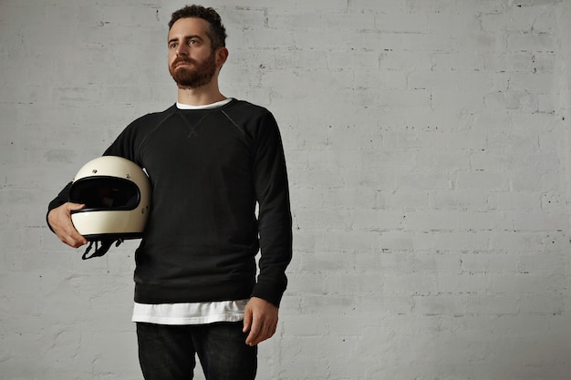 Bearded motorcyclist in black and white holding a blank white motorcycle helmet under his arm looking ahead setting