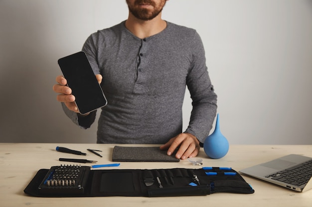 Free photo bearded professional shows repaired fixed smartphone after service replacement, above his specific tools in toolkit bag near laptop on wooden white table