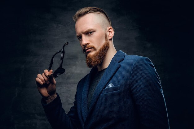 Bearded redhead male dressed in a blue suit and sunglasses over dark grey background.