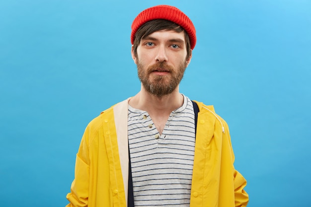 Free photo bearded sailor dressed in red hat and yellow anorak posing against blue wall. serious man with beard having blue charming eyes